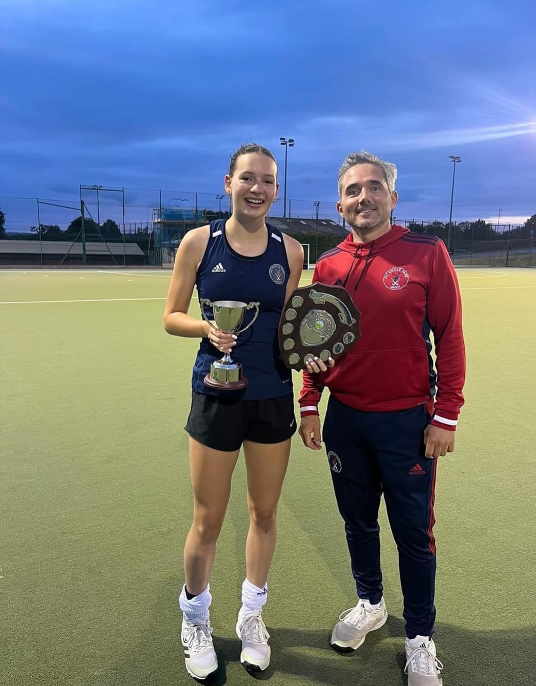 HC Knole Park player holding a cup and shield alongside her coach